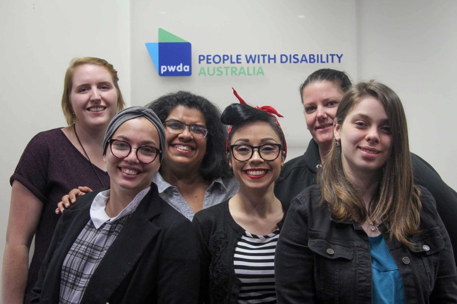- People with Disability Australia Group of six smiling people standing in front of a PWDA sign on the wall in the PWDA office.
