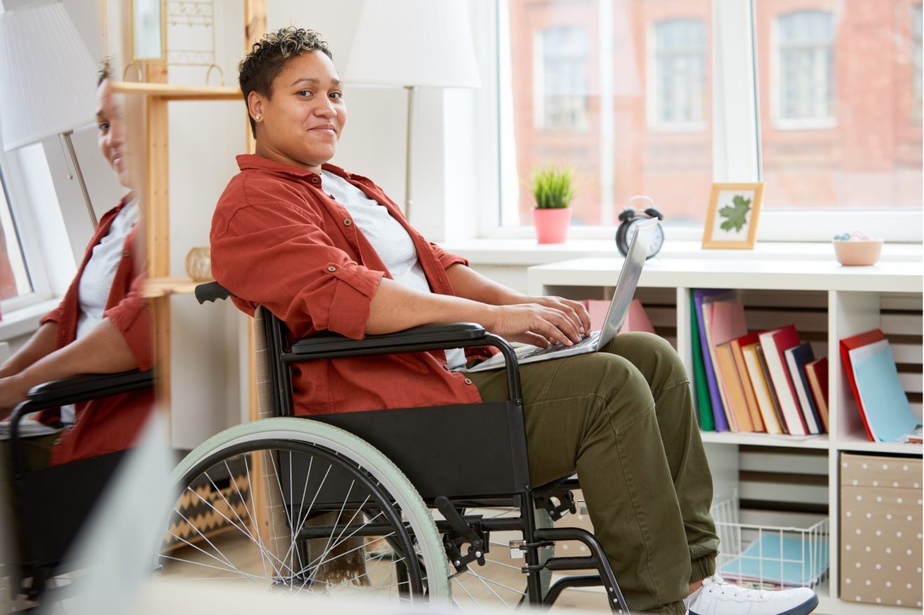 LanguageGuide image - People with Disability Australia A person viewed from side on, sitting in front of a large window in a wheelchair and smiling. There is a laptop resting on their lap, and they are wearing khaki pants, white t-shirt and a re shirt over the top.