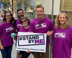 Five people standing on a city street, with one holding a white sign with a logo which says hashtag stand by me in a rectangle, with save disability advocacy in NSW written below it. They are all wearing purple t-shirts with the same logo.