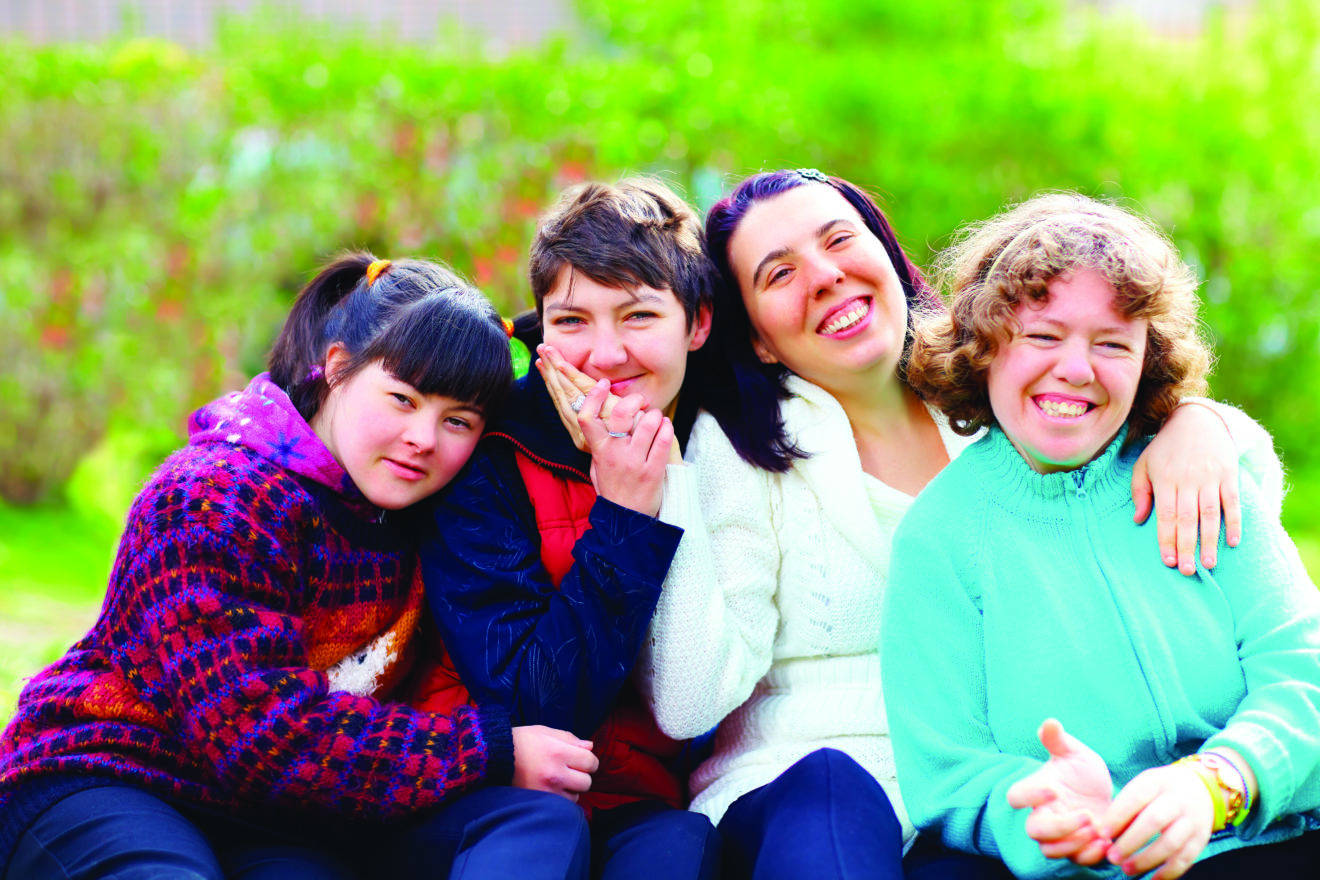 A group of women sitting together with their arms around each other. They are smiling at the camera. There are green bushes behind them.