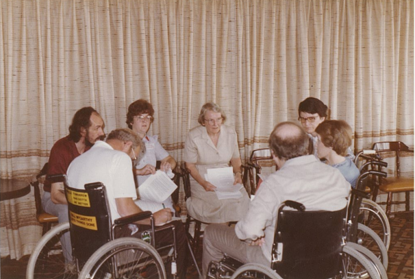 Possibly80s - People with Disability Australia Seven people, mostly wheelchair users, sitting in a close circle in discussion and holding paperwork. The photo was taken in the 1980’s.