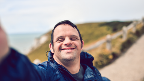 Man taking a selfie photo, as seen from his camera view, and smiling happily. In the background, a path can be seen which continues up the mountain he is on next to the ocean. - People with Disability Australia Man taking a selfie photo, as seen from his camera view, and smiling happily. In the background, a path can be seen which continues up the mountain he is on next to the ocean.