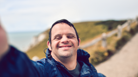 Man taking a selfie photo, as seen from his camera view, and smiling happily. In the background, a path can be seen which continues up the mountain he is on next to the ocean.