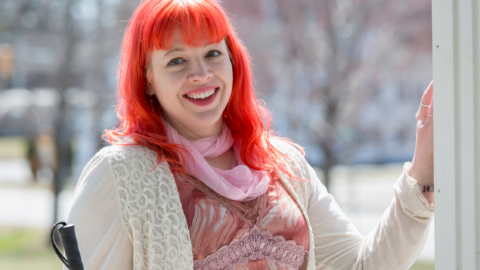 - People with Disability Australia Woman standing in a doorway that leads outside on a bright sunny day, with one hand resting on the frame and smiling happily. She has bright pinky red long hair, and is holding a white cane.