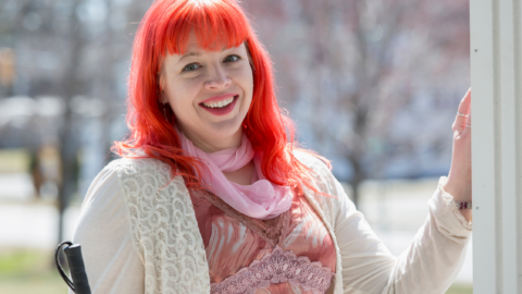 Woman standing in a doorway that leads outside on a bright sunny day, with one hand resting on the frame and smiling happily. She has bright pinky red long hair, and is holding a white cane.