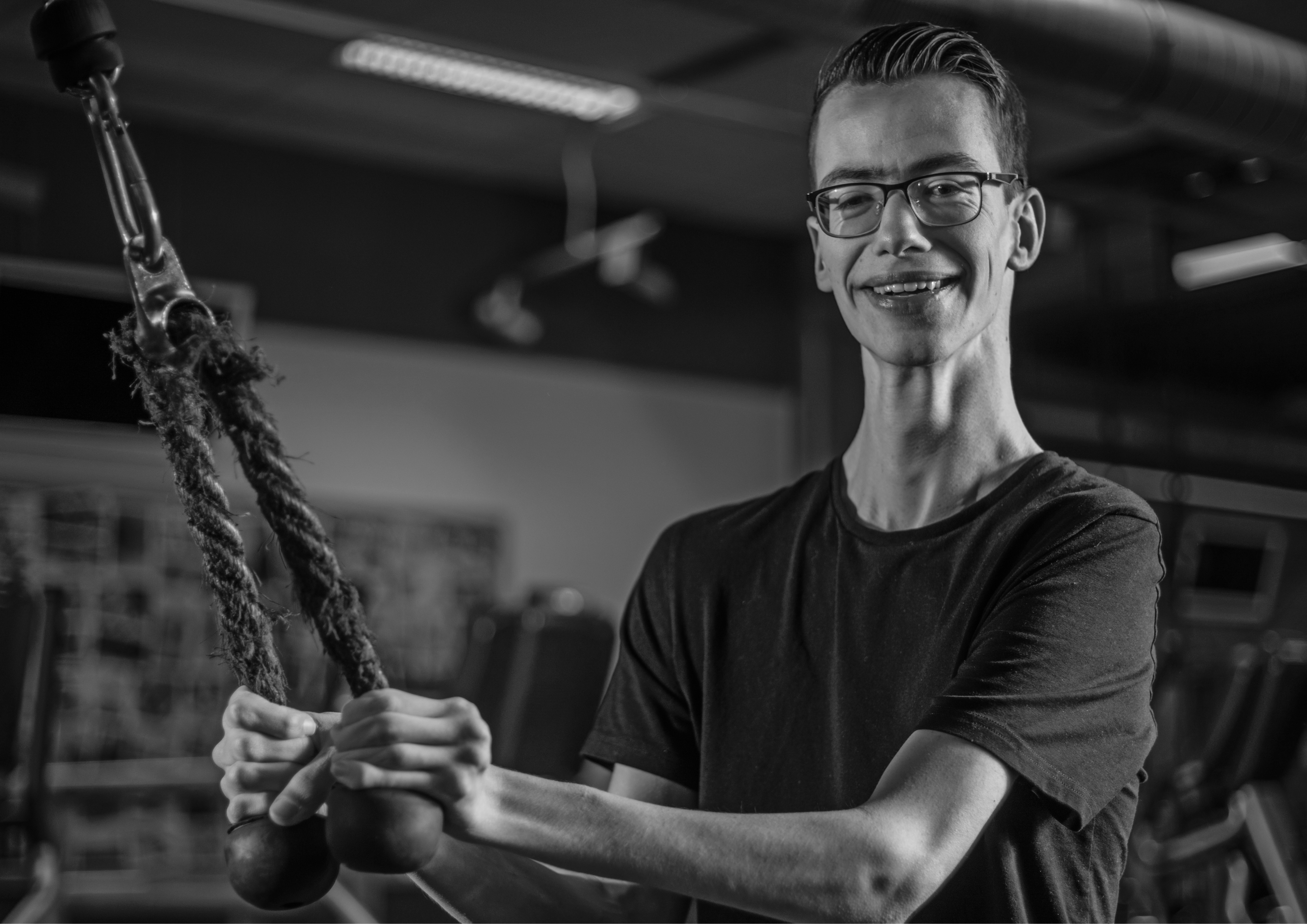 A person at a gym who is looking at the camera and smiling happily. He is holding onto equipment which has a rope attached to weights just out of shot.