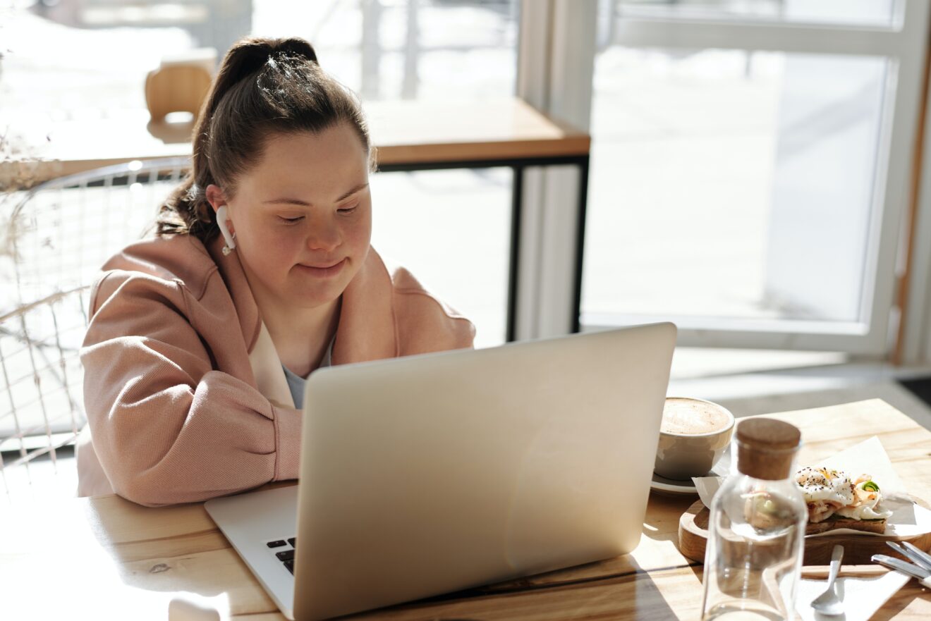 Woman sitting at a cafe table - People with Disability Australia Woman sitting at a cafe table with the sun streaming in behind her, she is looking at an open laptop in front of her, she has earbuds in her ears and coffee and food can be seen on the table beside her.