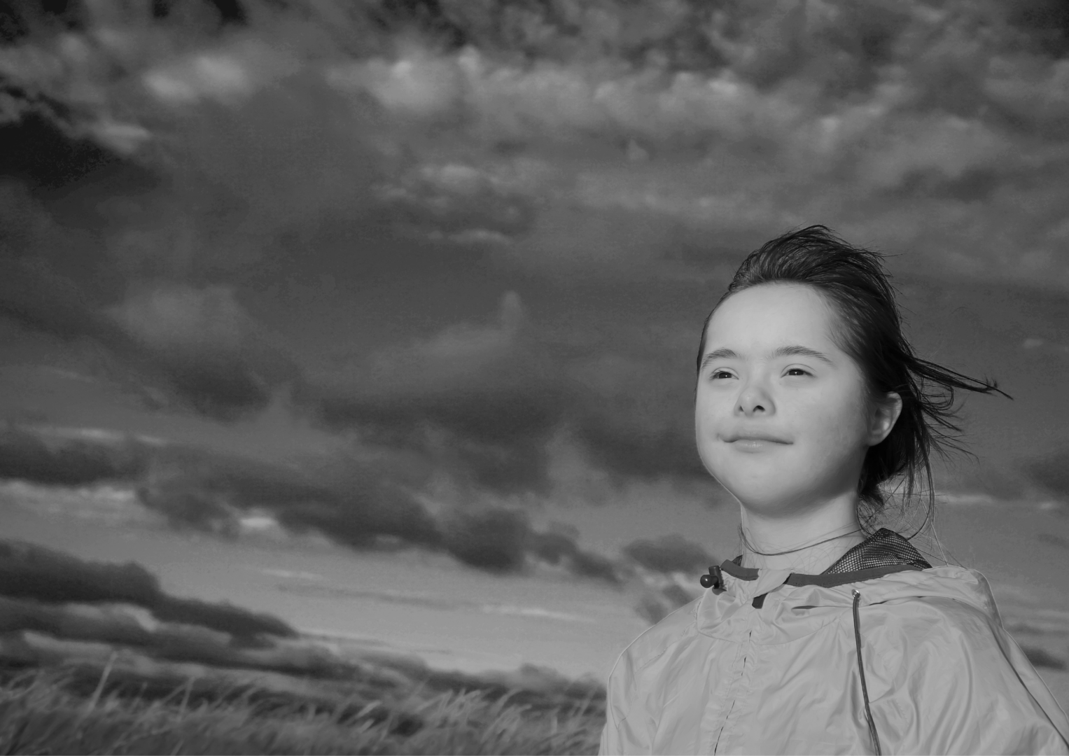 - People with Disability Australia Looking slightly up at a young person who is standing in a field and looking upwards with a content look on their face. A dramatic dark cloudy sky fills most of the frame.