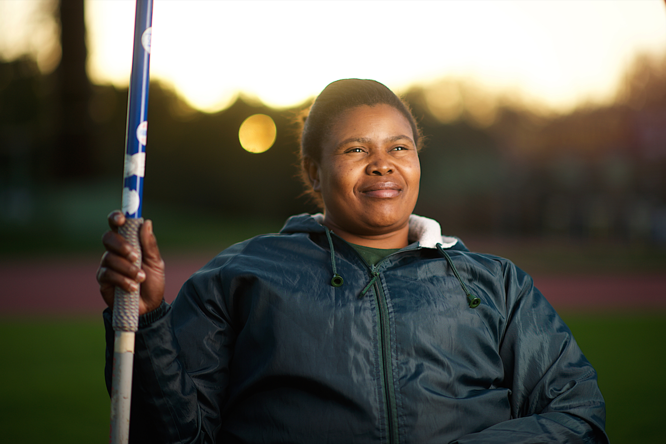 Javelin (2560 × 1708 px) - People with Disability Australia A person in a field with the sun behind them. They are smiling slightly and holding what appears to be a javelin.
