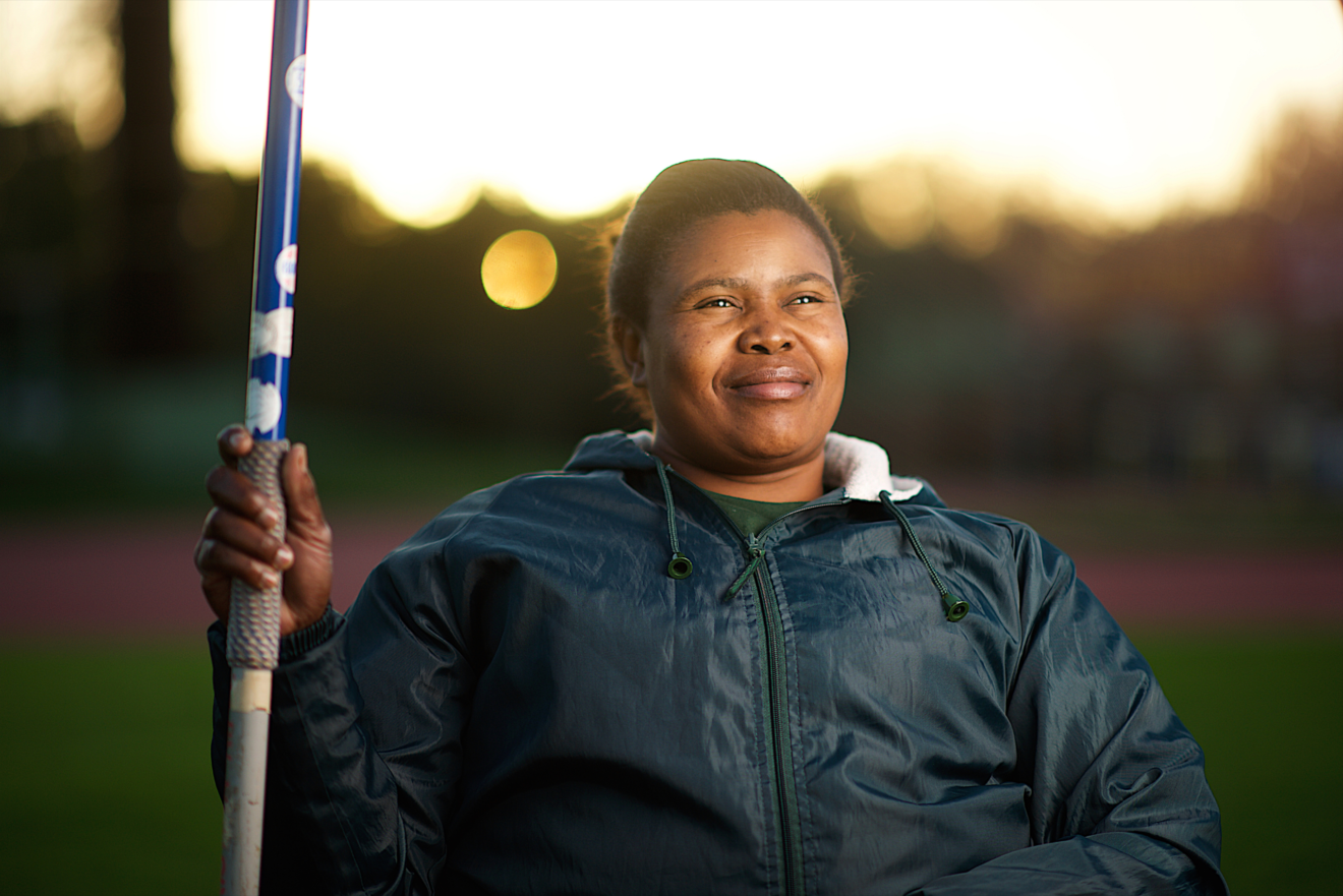 A person in a field with the sun behind them. They are smiling slightly and holding what appears to be a javelin.