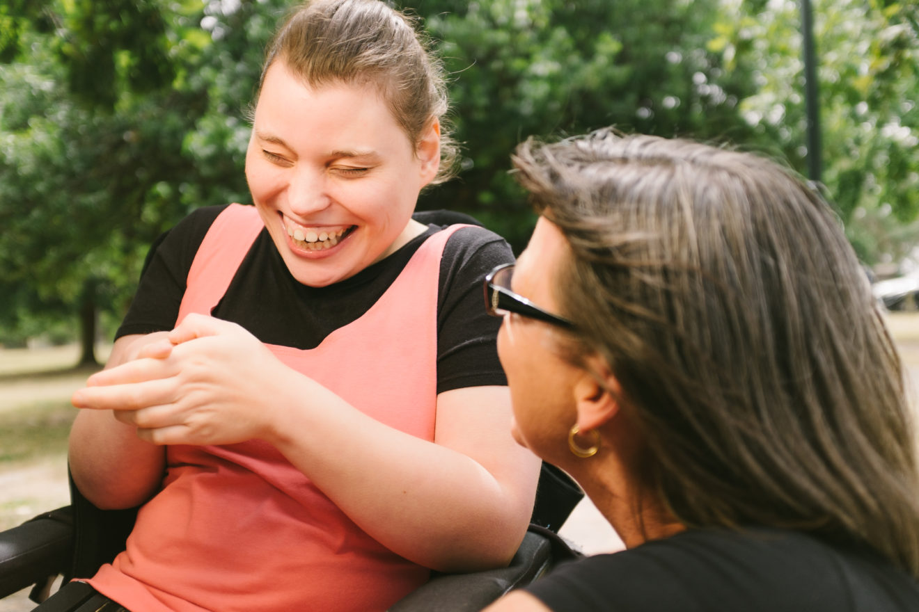- People with Disability Australia Mother interacting with her daughter in a park, she is leaning in close and tucking her daughters hair behind her ear. The daughter is looking down and smiling.