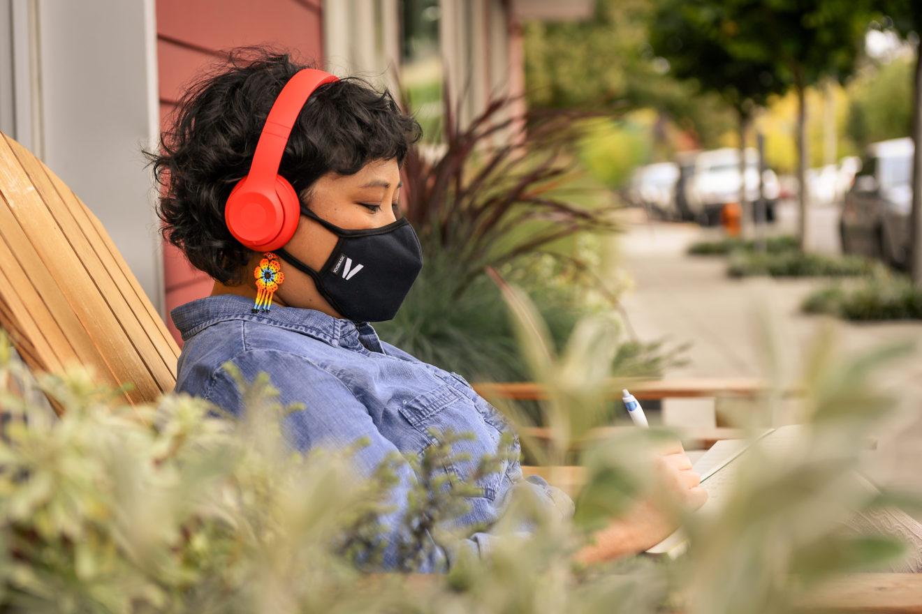 - People with Disability Australia Person viewed from side on and from behind some plants, they are wearing a face mask and headphones and sitting outside a cafe on a deck chair and writing in a notebook.