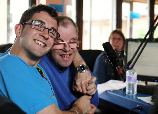 - People with Disability Australia Two people smiling in a studio and hugging each other. There is a microphone in front of them.