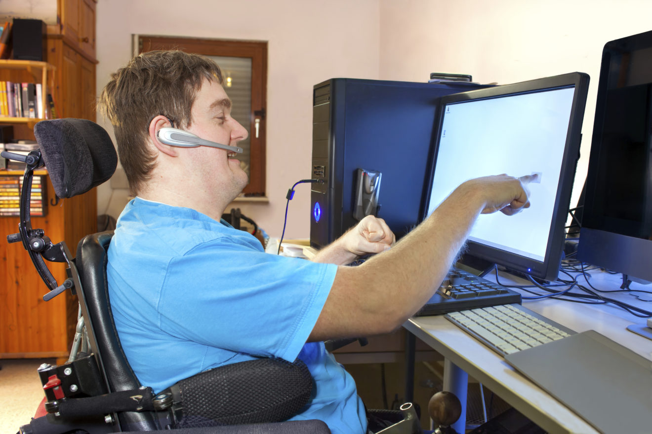 - People with Disability Australia Man with disability sitting in multifunctional wheelchair, using a computer with a wireless headset, reaching out to touch the touch screen.