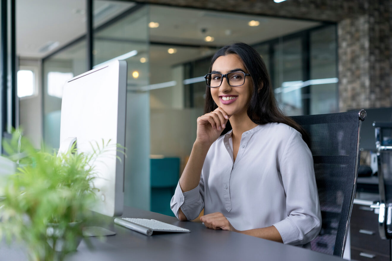 Portrait of cheerful young businesswoman wearing glasses sitting at her workplace with computer in office looking at camera with hand on chin and smiling