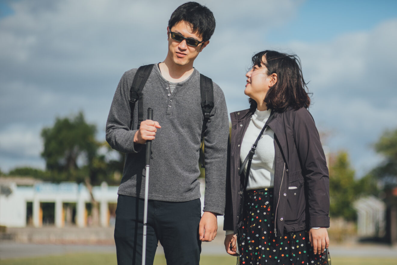 Two people standing closely together outside on a partly cloudy day, the person on the left is holding a white cane and is wearing glasses - People with Disability Australia Two people standing closely together outside on a partly cloudy day, the person on the left is holding a white cane and is wearing glasses, the other person is looking up at him and smiling